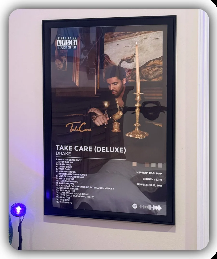 A framed Take Care (Deluxe) album print by Drake, with its track list and a Spotify code, hanging on a pale wall lit by a small purple lamp.